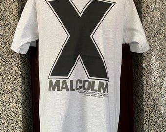 May include: A light gray t-shirt featuring a large black "X" graphic with a white outline. Below the "X" is the name "MALCOLM" in gray block letters. The shirt is displayed on a mannequin.