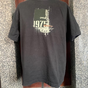 Puede incluir: Camiseta negra con un diseño gráfico que presenta el texto "1975" y la marca "Oakley". El diseño incluye una forma rectangular con un aspecto descolorido y desgastado. La camiseta se muestra en un maniquí.