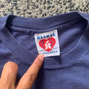 May include: Close-up of a navy blue t-shirt with a white tag. The tag reads "HECKEL" in blue letters, with a red heart logo. Below the logo, the text "TRADE MARK IYUGAOKA" is printed.