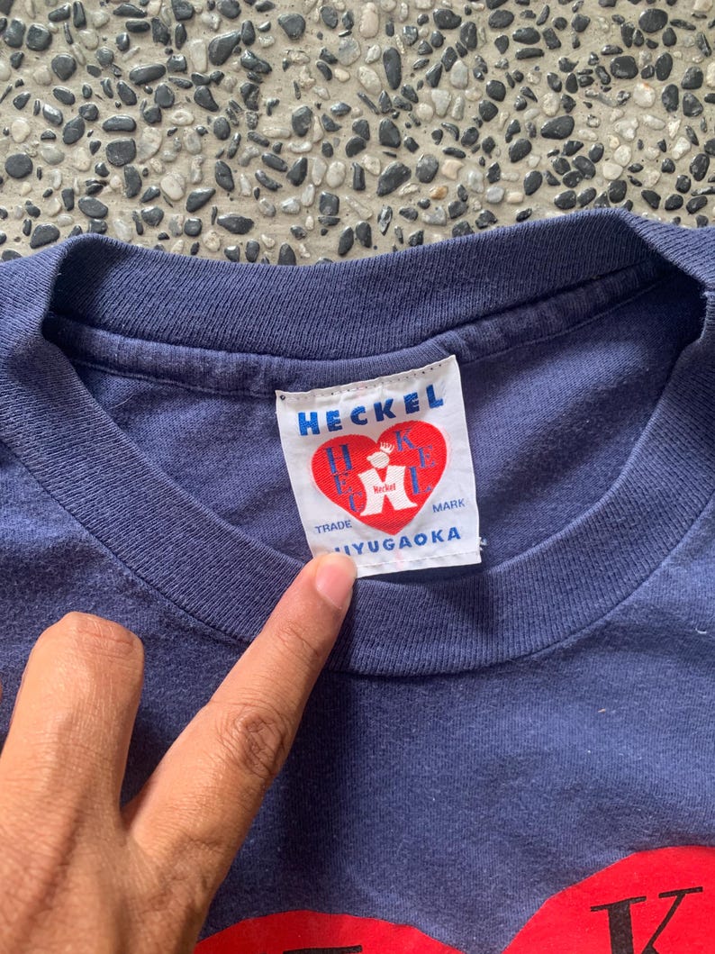 May include: Close-up of a navy blue t-shirt with a white tag. The tag reads "HECKEL" in blue letters, with a red heart logo. Below the logo, the text "TRADE MARK IYUGAOKA" is printed.