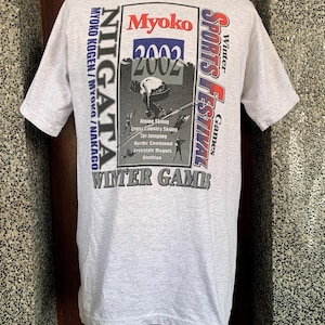 Puede incluir: Camiseta gris claro con un gráfico que promociona el Festival de Deportes de Invierno Myoko 2002. El diseño incluye texto como "NIIGATA", "Myoko" y "Winter Games", junto con ilustraciones de deportes de invierno.