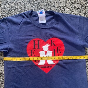 May include: Navy blue t-shirt with a red heart graphic. The heart contains the letters "H K E I E" and a crown. The text "JIYUGAOKA" is below the heart. The shirt is approximately 23 inches wide.