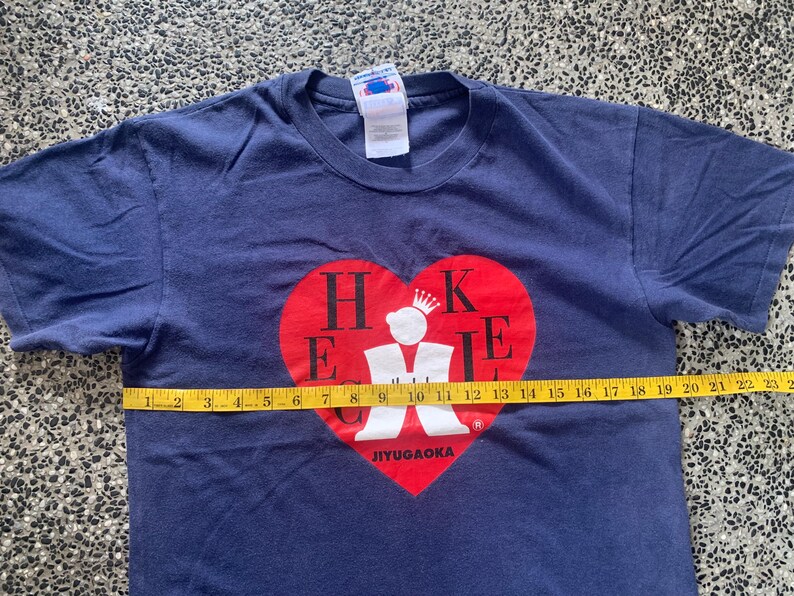 May include: Navy blue t-shirt with a red heart graphic. The heart contains the letters "H K E I E" and a crown. The text "JIYUGAOKA" is below the heart. The shirt is approximately 23 inches wide.