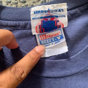 May include: Close-up of a navy blue Hanes Beefy-T adult medium t-shirt. The tag shows the brand name and size, along with the text "100% preshrunk cotton". The shirt's fabric is made in the USA.
