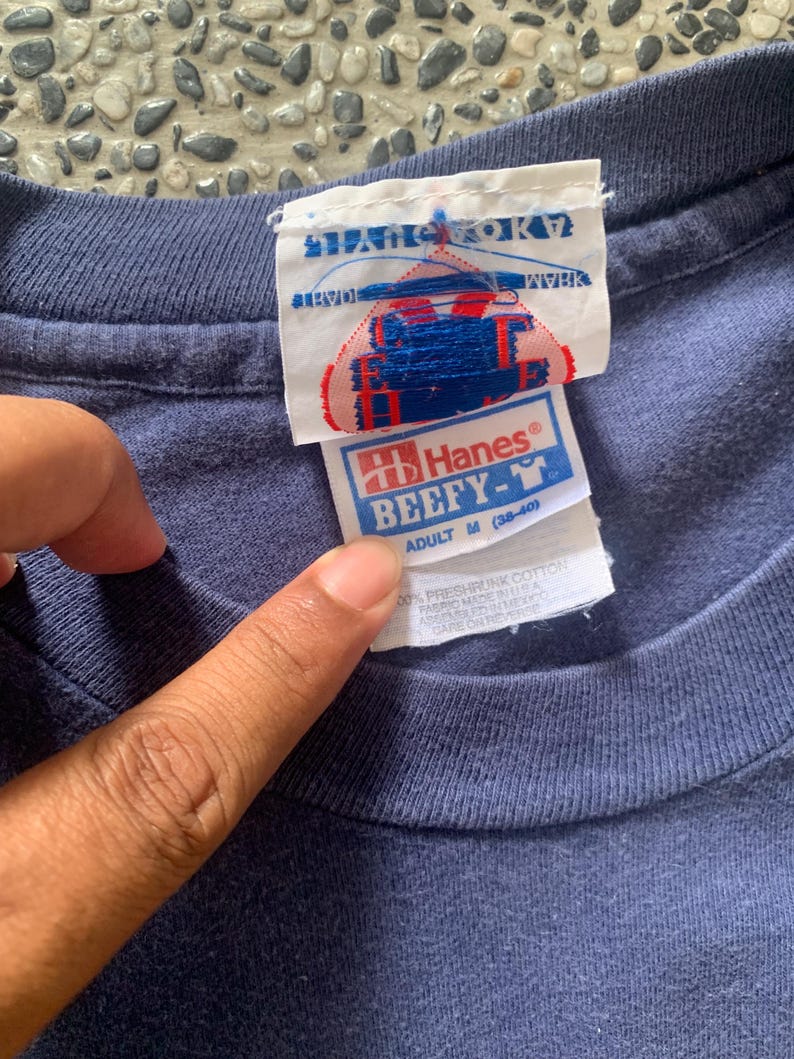 May include: Close-up of a navy blue Hanes Beefy-T adult medium t-shirt. The tag shows the brand name and size, along with the text "100% preshrunk cotton". The shirt's fabric is made in the USA.
