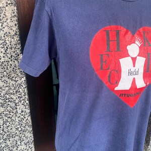 May include: Navy blue t-shirt featuring a large red heart graphic with the letters HECK and a white figure wearing a crown. The text "JIYUGAOKA" is below the heart. The shirt is made of a soft material.
