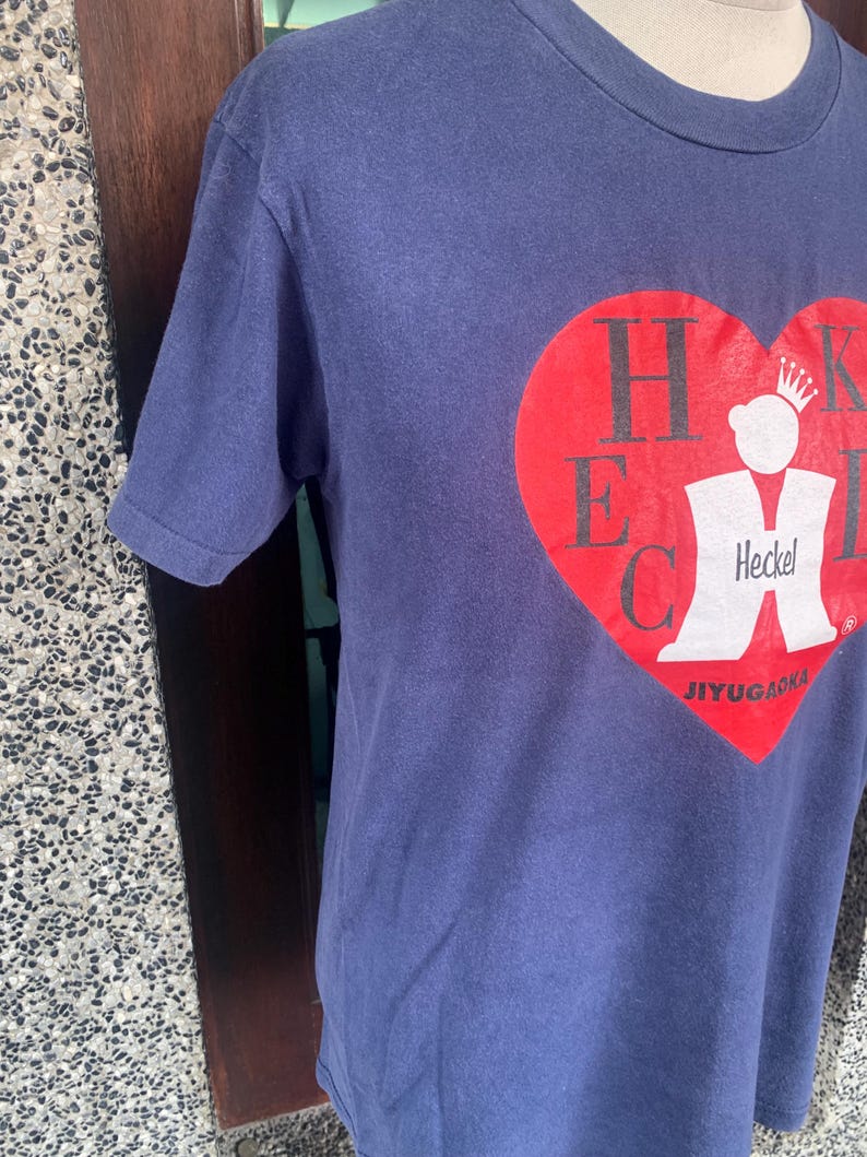 May include: Navy blue t-shirt featuring a large red heart graphic with the letters HECK and a white figure wearing a crown. The text "JIYUGAOKA" is below the heart. The shirt is made of a soft material.