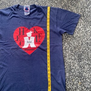 May include: Navy blue t-shirt with a red heart graphic containing the name "Heckel" and the text "JIYUGAOKA". The shirt is approximately 28 inches long, measured with a yellow tape measure. The shirt has short sleeves and a crew neck.