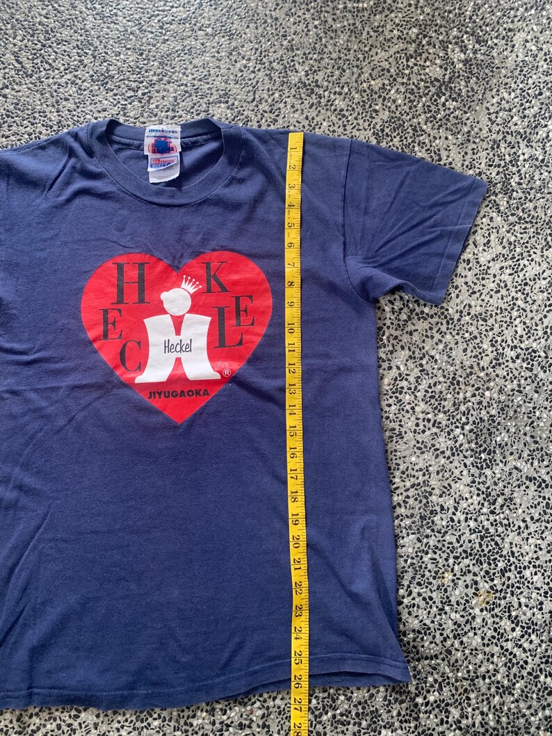 May include: Navy blue t-shirt with a red heart graphic containing the name "Heckel" and the text "JIYUGAOKA". The shirt is approximately 28 inches long, measured with a yellow tape measure. The shirt has short sleeves and a crew neck.