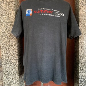 Puede incluir: Camiseta negra de manga corta con el texto "Intel Pentium 4 Processor Summer 2003 Championship". La camiseta también presenta un logotipo de "Intel Inside Pentium 4" en el pecho izquierdo.
