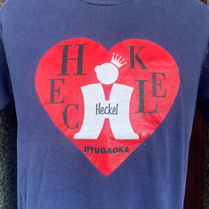 May include: Navy blue t-shirt featuring a red heart-shaped graphic with the word "Heckel" and a crown. The graphic also includes the text "JIYUGAOKA". The shirt is made of a soft material.