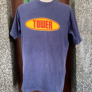 Peut inclure: Un t-shirt bleu vintage avec un logo ovale jaune sur lequel est inscrit "TOWER" en lettres rouges.