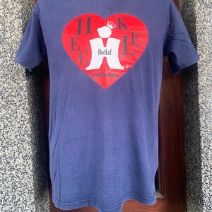 May include: A navy blue t-shirt featuring a red heart-shaped graphic with the word "HECKEL" in white letters. The graphic also includes a white figure with a crown and the text "BYUGONA". The shirt is displayed on a mannequin.