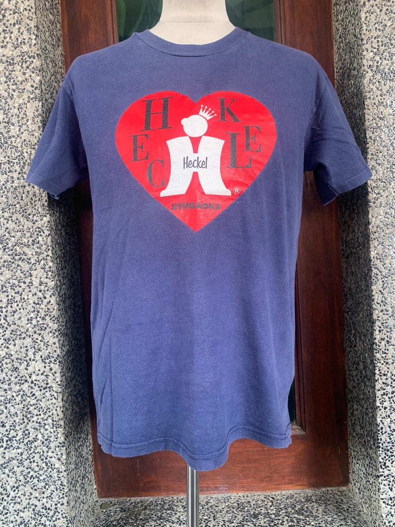 May include: A navy blue t-shirt featuring a red heart-shaped graphic with the word "HECKEL" in white letters. The graphic also includes a white figure with a crown and the text "BYUGONA". The shirt is displayed on a mannequin.