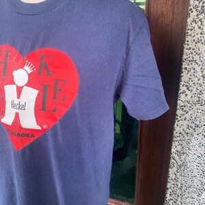 May include: Navy blue t-shirt featuring a large red heart design with the text "Heckel" and "Yugaoaka" in white. The shirt has a crew neck and short sleeves. The design includes a crown.