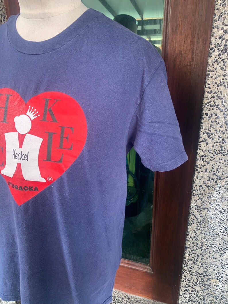 May include: Navy blue t-shirt featuring a large red heart design with the text "Heckel" and "Yugaoaka" in white. The shirt has a crew neck and short sleeves. The design includes a crown.