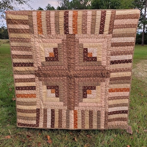 May include: A square quilt featuring a log cabin block pattern in shades of brown, tan, cream, and orange. The center displays a cross motif surrounded by vertical striped borders made of various patterned fabric strips with visible stitching.