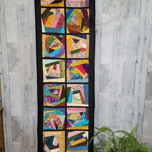 May include: A long, black fabric wall hanging with 12 colorful squares. Each square is made of different patterned fabrics in shades of pink, yellow, blue, green, red, and brown. The squares are arranged in a vertical row.