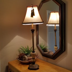 May include: A table lamp with a cream-colored shade and gold tassel accents sits on a wooden table next to a decorative mirror. The lamp is on and casting a warm glow.