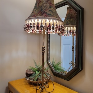 May include: A gold table lamp with a floral patterned lampshade and a fringe trim. The lamp is on a wooden table with a green plant and a brown wicker basket.