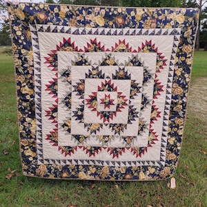 May include: A square patchwork quilt featuring a central star motif surrounded by concentric geometric patterns in cream, navy blue, and deep red. The border displays a floral print with yellow roses on a dark navy background.