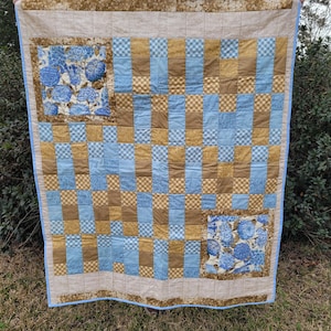 May include: A handmade quilt with a patchwork design. The quilt features squares of blue, brown, and patterned fabric. Two large squares with blue floral patterns are set in the corners. The quilt is trimmed with a light brown border and a blue binding.