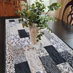 May include: A rectangular table runner featuring a patchwork design of black, white, and gray fabrics with various patterns including swirls, dots, and floral motifs. A clear glass vase filled with green eucalyptus stems sits on top.