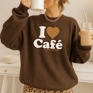 May include: A brown sweatshirt with the text "I ♥ Café con leche" in white and a brown heart. The sweatshirt is paired with a gold chain necklace and a leopard print skirt. A white mug and a glass jar with a straw are also visible.