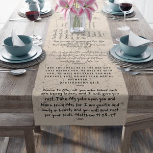 May include: Beige table runner with printed scriptural text and a map design. The runner is set on a wooden table with place settings, wine glasses, and a vase of pink flowers. The text includes Bible verses.