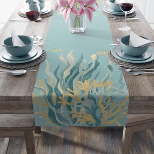May include: A light blue table runner with an underwater design featuring seaweed and coral in shades of gold and teal. The runner is set on a wooden table with place settings, wine glasses, and a vase of pink flowers.