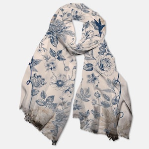 May include: A cream-colored scarf with a blue floral pattern. The design features various flowers, leaves, and a hummingbird. The scarf has a fringed edge and is draped, showcasing the intricate botanical print.
