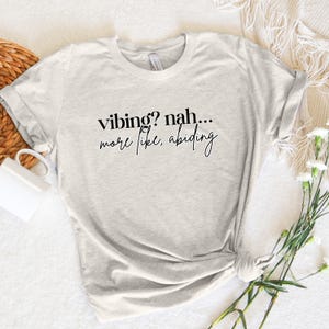 Puede incluir: Una camiseta gris claro con texto negro que dice "vibing? nah... more like, abiding".