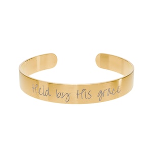 Può includere: Un bracciale a polsino color oro con l'iscrizione "Held by His grace" in stile scritto a mano. Il bracciale ha una finitura liscia e lucida e un design semplice e aperto. Questo gioiello è adatto all'uso quotidiano.
