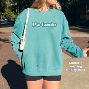 Peut inclure: Un sweat-shirt ras du cou turquoise avec le texte "Pa'lante onward" en bleu clair. Le sweat-shirt est de couleur unie et a des manches longues. Le modèle porte un sweat-shirt de couleur "Chalky mint".