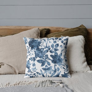 May include: A blue and white floral patterned throw pillow on a bed with a grey and white blanket. The pillow is on top of a tan pillow and a green pillow.