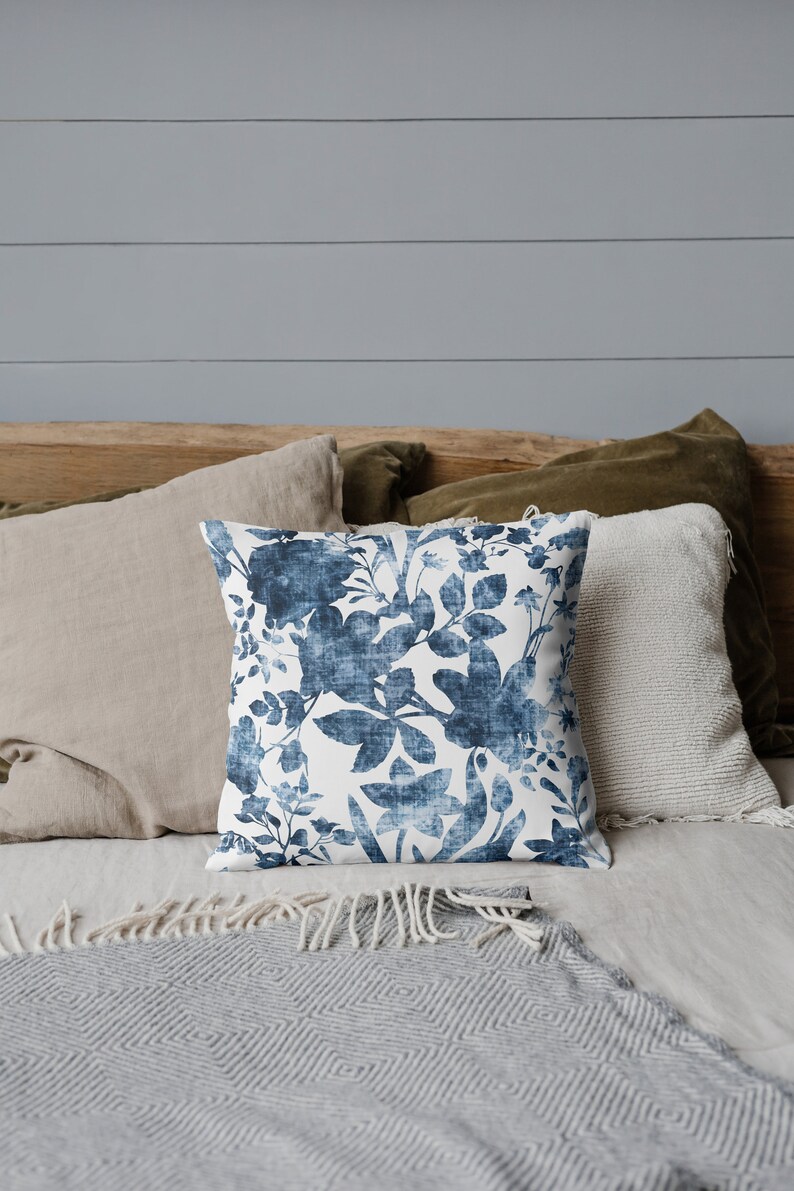 May include: A blue and white floral patterned throw pillow on a bed with a grey and white blanket. The pillow is on top of a tan pillow and a green pillow.