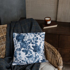 May include: A wicker chair with a blue and white floral patterned cushion, a grey and yellow checked throw blanket, and a white cushion.