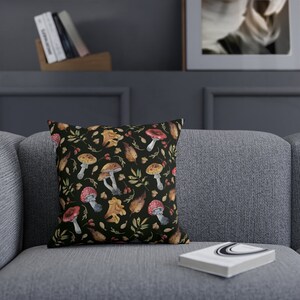 May include: A black throw pillow with a colorful mushroom and leaf pattern. The pillow is on a gray couch with a book on the couch.