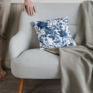 May include: A light grey sofa with a throw blanket and a blue and white floral cushion. The sofa has wooden legs and is sitting on a wooden floor.
