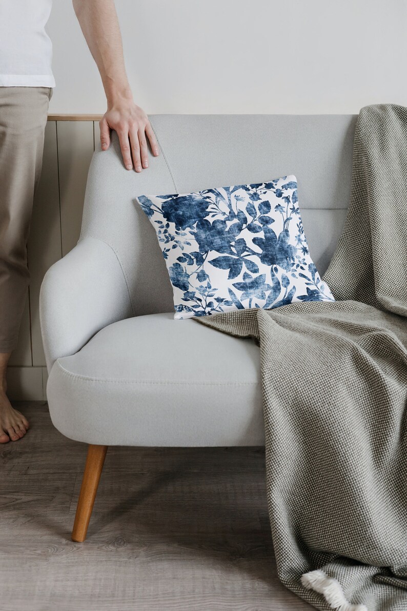May include: A light grey sofa with a throw blanket and a blue and white floral cushion. The sofa has wooden legs and is sitting on a wooden floor.