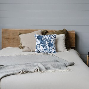 May include: A bed with a white duvet and a grey throw blanket. The bed has a wooden headboard and is decorated with pillows, including one with a blue floral pattern.