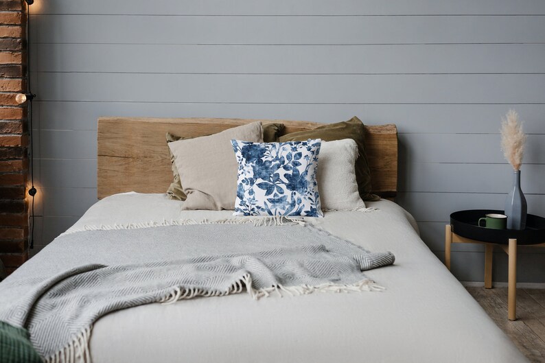 May include: A bed with a white duvet and a grey throw blanket. The bed has a wooden headboard and is decorated with pillows, including one with a blue floral pattern.