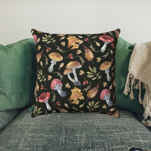 May include: A black throw pillow with a watercolor pattern of mushrooms, leaves, and berries. The pillow is on a green couch with a white throw blanket.