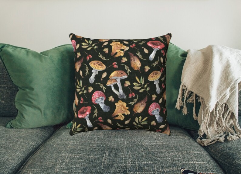 May include: A black throw pillow with a watercolor pattern of mushrooms, leaves, and berries. The pillow is on a green couch with a white throw blanket.