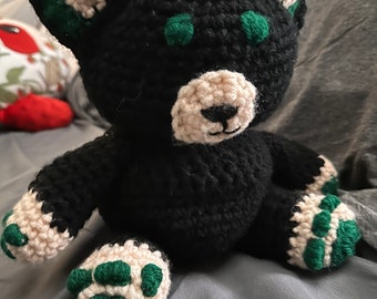 May include: A black crocheted cat plush toy with green ears and paws. The cat has a white face and a pink nose.