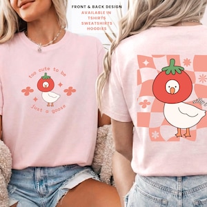 May include: Light pink t-shirt featuring a cartoon goose wearing a tomato hat. The front design reads "too cute to be just a goose." The back has the same goose design with the words "silly goose" and a checkered pattern.