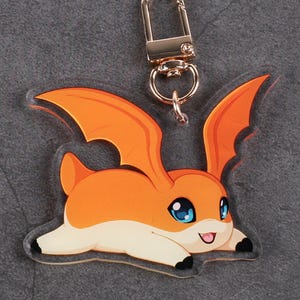 May include: An acrylic keychain featuring a cartoon creature with orange fur, large blue eyes, and wings. The character is in a prone position with a small tongue visible. It is attached to a gold-toned metal lobster clasp hardware.