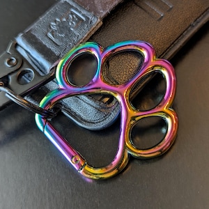 Op de afbeelding: Een regenboogkleurige metalen sleutelhanger in de vorm van een pootafdruk met een karabijnhaak bevestigd aan een zwarte sleutelhouder van kunstleer. De pootafdruk heeft een verloop van iriserende kleuren waaronder paars, blauw, groen, geel en oranje.