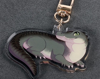 Könnte beinhalten: Ein klarer Acryl-Schlüsselanhänger mit einem Cartoon-Alligator in Grau-, Lila- und Weißtönen. Der Alligator hat grüne Augen und einen goldfarbenen Verschluss. Der Schlüsselanhänger ist zum Anbringen an Schlüsseln oder einer Tasche gedacht.