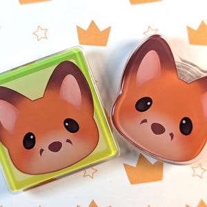 Peut inclure: Deux accessoires sur le thème du renard. L'un est carré avec un bord vert, l'autre en forme de renard. Les deux présentent un visage de renard de dessin animé avec une fourrure orange, des yeux sombres et un petit nez. Le fond a un motif de couronnes et d'étoiles.